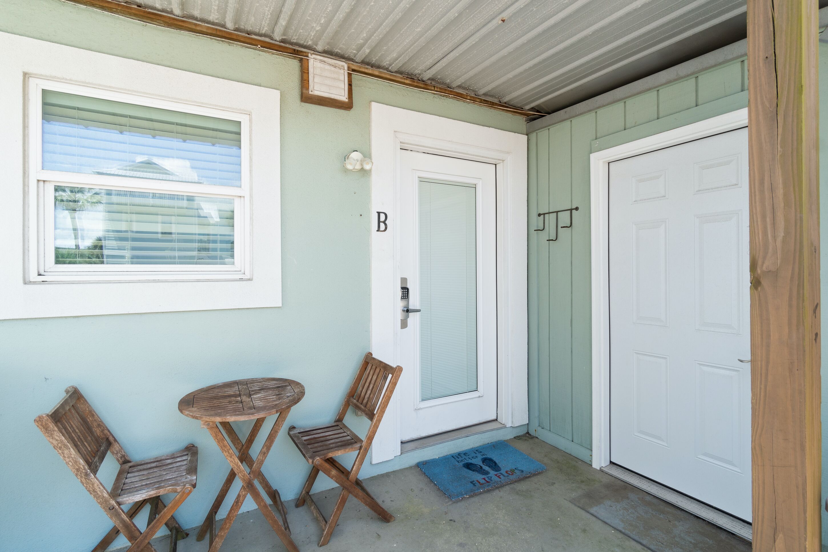 A small porch with two wooden chairs and a table, a white door with a 'B' sign, and a blue mat on the floor.