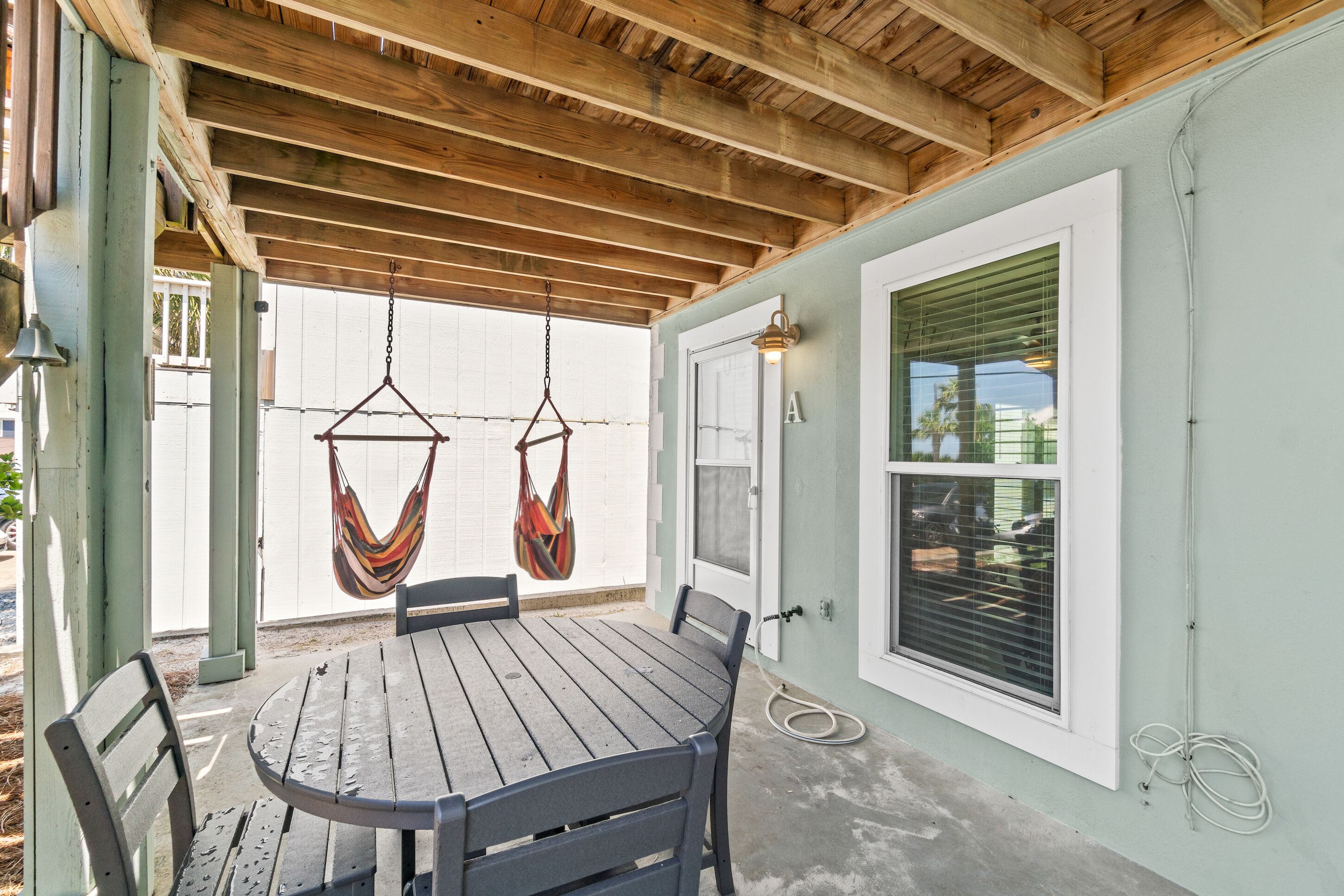 A covered patio with a wooden table and chairs, a hanging hammock, and a door with a window.