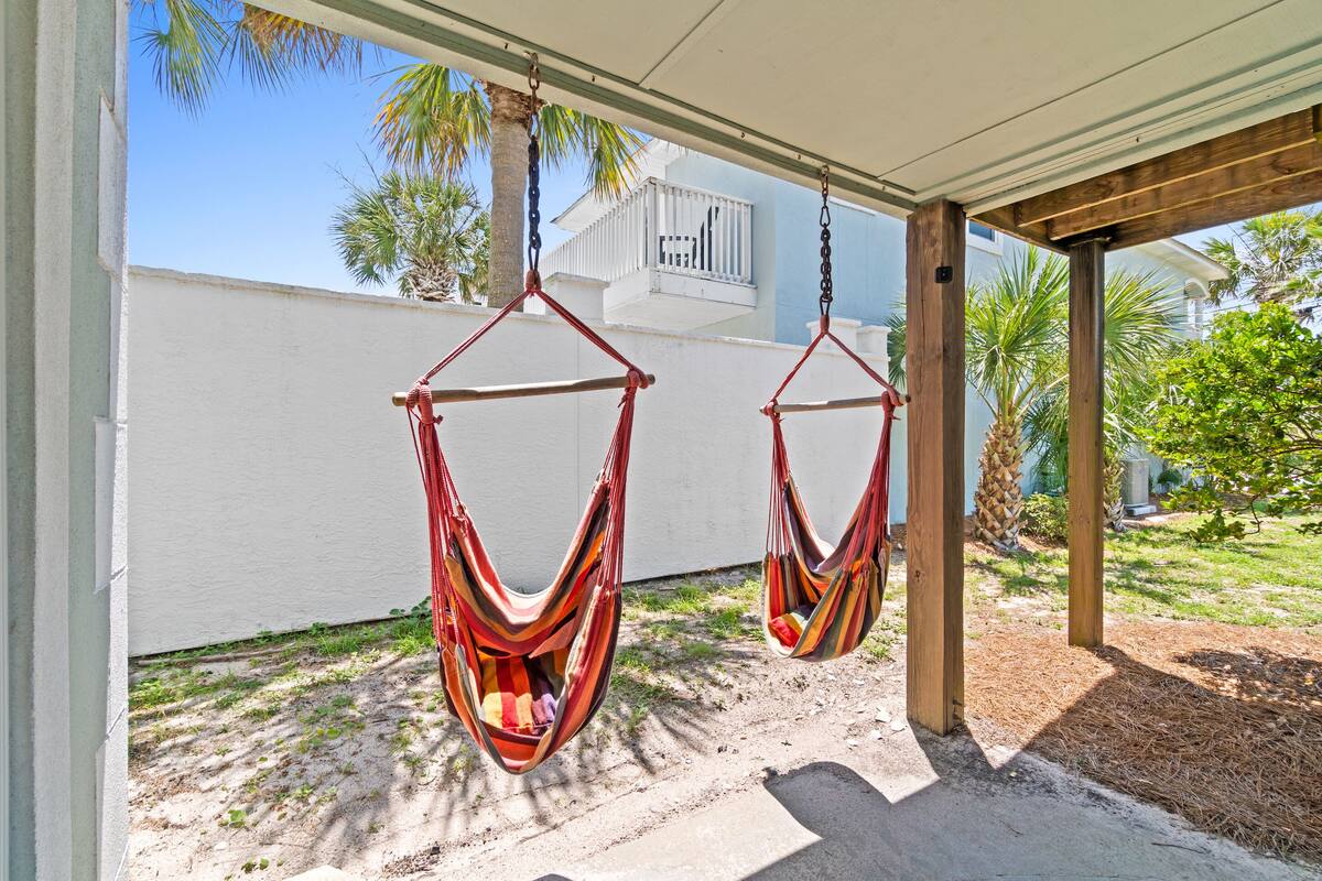 Dos sillas hamaca colgadas bajo una pérgola de madera, con una pared blanca y una palmera al fondo.