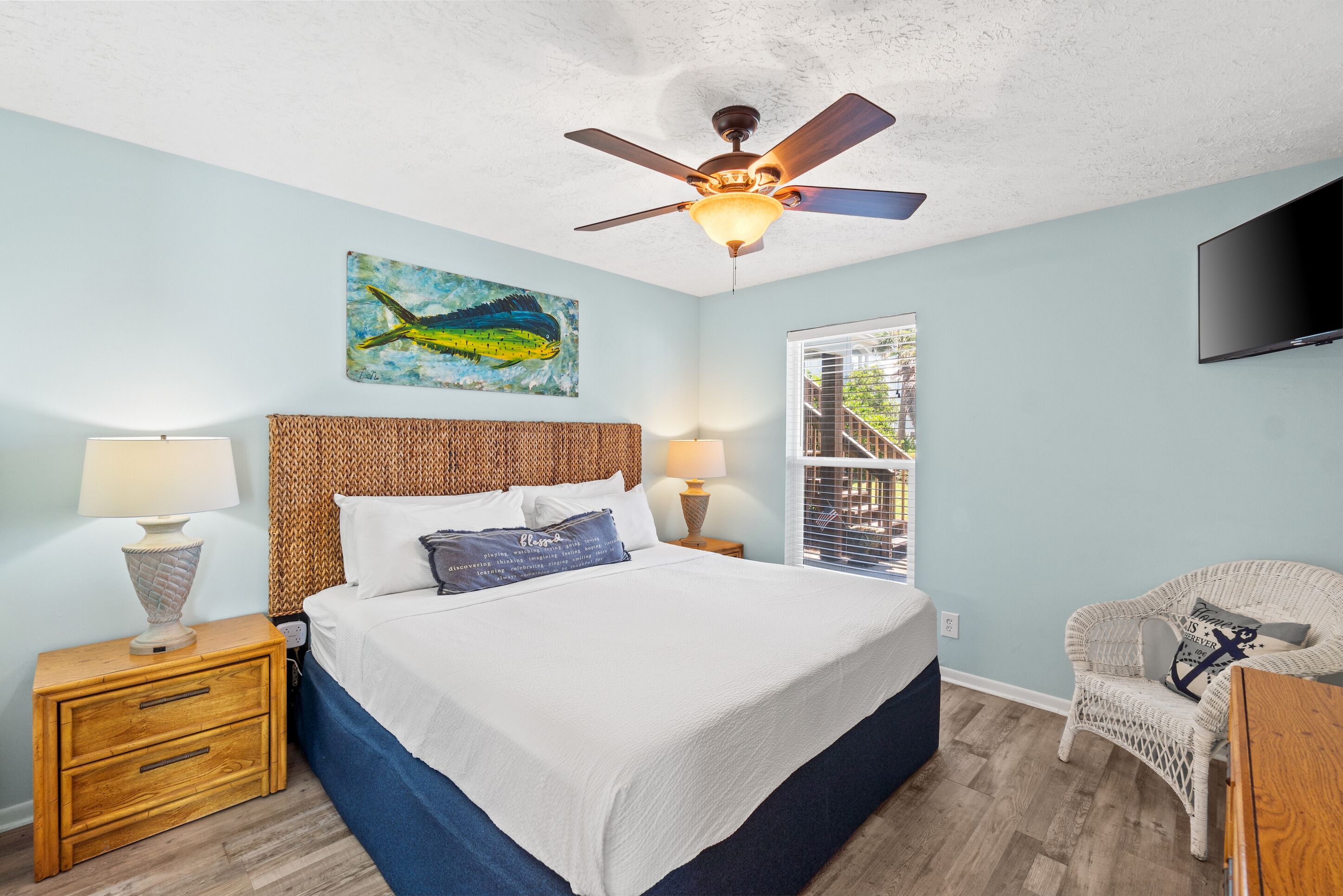 A bedroom with a bed, wooden nightstands, a ceiling fan, a painting, a window, and a TV.