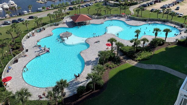 A large, multi-pond swimming area with a surrounding deck, palm trees, and a marina in the background. - Yacht Club Villas #2-401 Relaxing Waterway View in Barefoot Resort! (North Myrtle Beach)