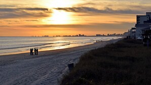 A beach with a sunset, people walking, and buildings in the background. - Yacht Club Villas #2-401 Relaxing Waterway View in Barefoot Resort! (North Myrtle Beach)