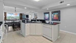 A modern kitchen with white cabinetry, a black countertop, and a dining area with a table and chairs. - Yacht Club Villas #2-401 Relaxing Waterway View in Barefoot Resort! (North Myrtle Beach)