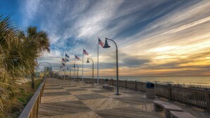 A wooden boardwalk with benches and street lamps, overlooking a body of water with flags and a Ferris wheel in the background. - Yacht Club Villas #2-401 Relaxing Waterway View in Barefoot Resort! (North Myrtle Beach)