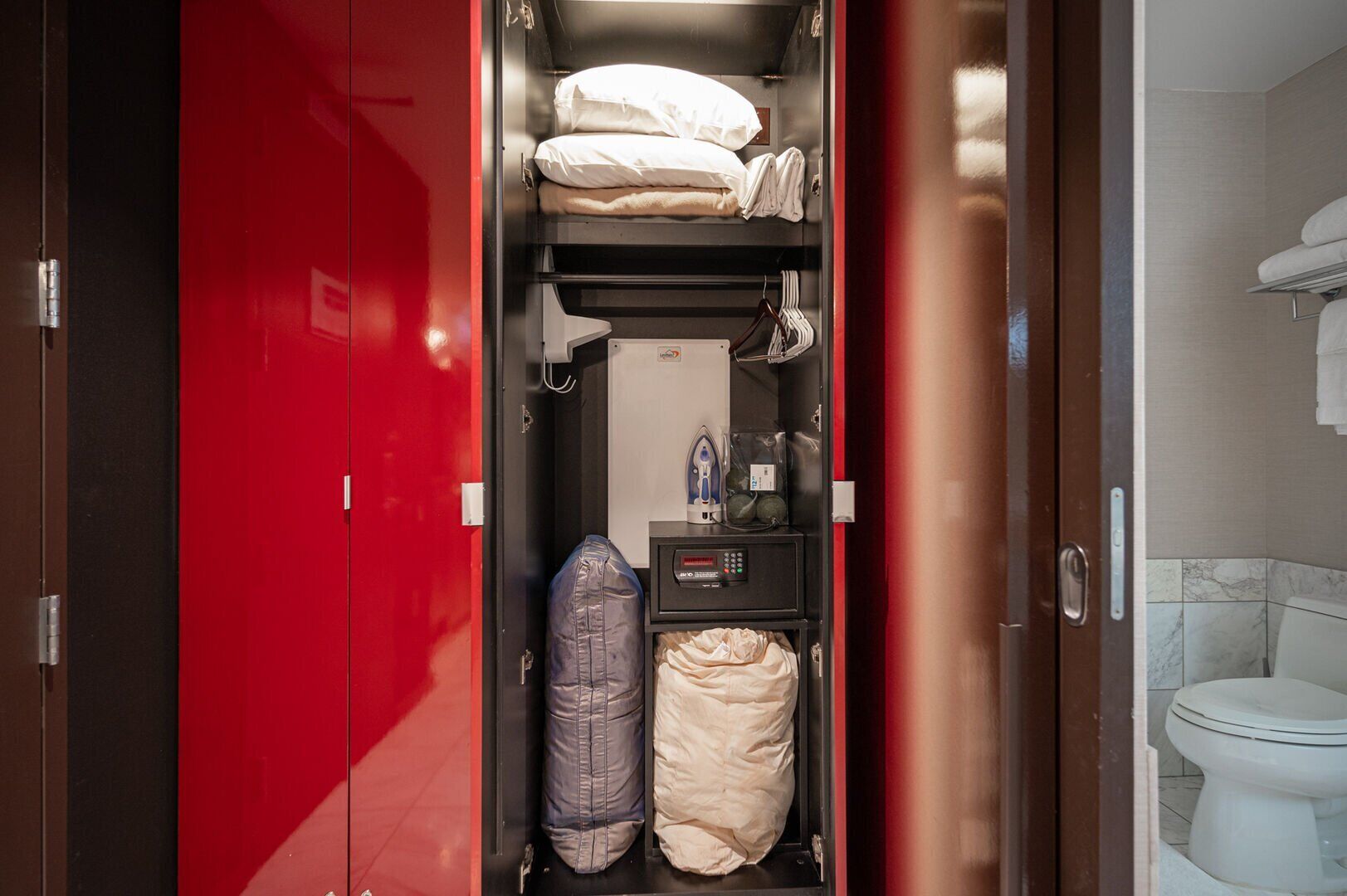 Een compacte hotelkamer met een rode garderobe, een bed met witte lakens, een klein strijkplankje en een toilet.