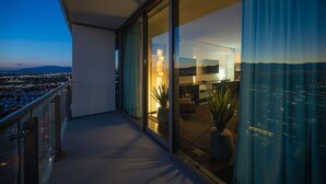 Uma varanda com vista para a cidade ao entardecer. No interior, há uma sala de estar moderna com uma televisão, um sofá e uma mesa de centro.