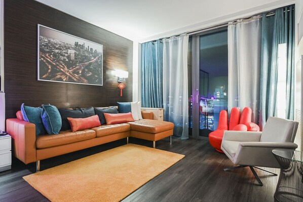 Un salon moderne avec un canapé d’angle, un fauteuil rouge et une petite table. Une grande fenêtre, ornée de rideaux, offre une vue sur le paysage urbain la nuit.