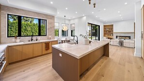 A modern kitchen with a central island, wooden cabinets, and large windows offering a view of greenery. - Genesta Villa - A Contemporary Modern Villa w/ Pool, Spa & Theater (Encino)