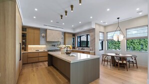 A modern kitchen with wooden cabinets, a central island, and a dining area with a table and chairs. - Genesta Villa - A Contemporary Modern Villa w/ Pool, Spa & Theater (Encino)