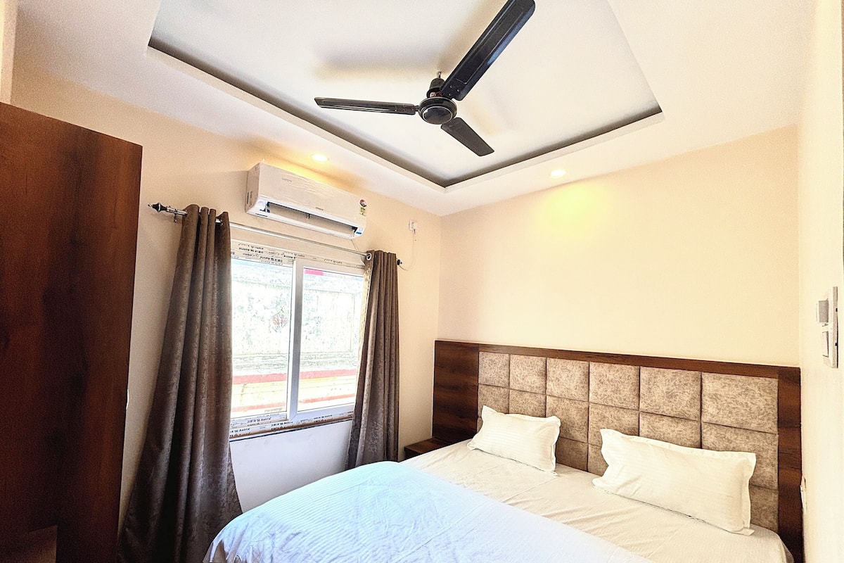 Habitación de hotel con cama, ventilador de techo, equipo de aire acondicionado y ventana con cortinas.