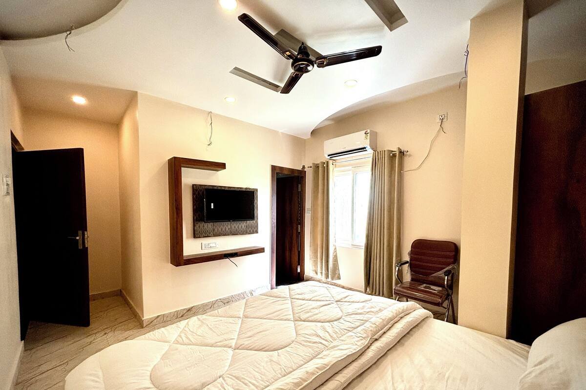 Un dormitorio con cama, televisor montado en la pared, ventilador de techo y ventana con cortinas.