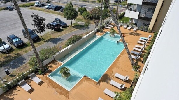 A rectangular swimming pool with a sandy deck and lounge chairs, surrounded by trees and a parking area.