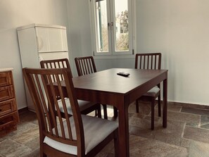 A dining area with a wooden table and chairs, a white refrigerator, and a window with a view of trees and buildings. - Tinos 4 Person House in Heart of Tinos Island (Tinos)