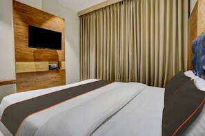 A hotel room with a bed, a flat-screen TV mounted on a wooden panel, and a large window with beige curtains.