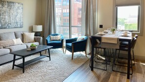 Una sala de estar moderna con sofá, sillón, mesa de comedor y un amplio ventanal con cortinas.