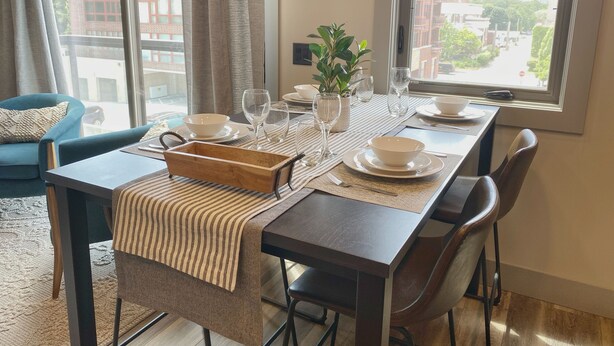 Comedor con una mesa dispuesta para cuatro, incluyendo platos, tazones, vasos y cubiertos. Hay una planta en la mesa y la habitación tiene ventanas amplias con vistas a edificios.