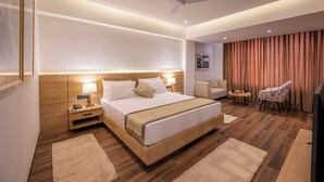 Une chambre d’hôtel moderne dotée d’un grand lit, de tables de chevet, d’un ventilateur de plafond, d’une télévision et d’un coin salon avec un fauteuil et une petite table.