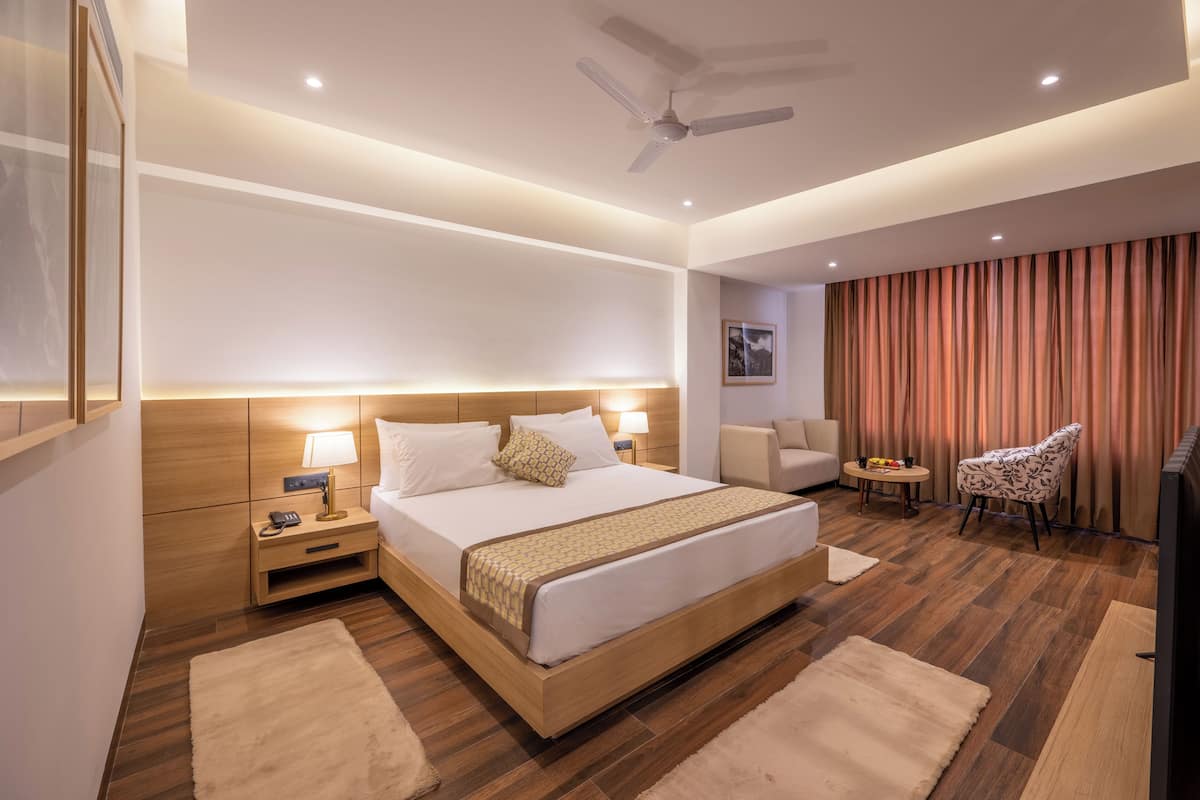 A modern hotel room with a large bed, bedside tables, a ceiling fan, a TV, and a seating area with a chair and a small table.