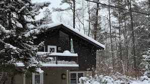 Une cabane en bois dotée d’un balcon, entourée d’arbres couverts de neige et bordée d’une grande pile de neige à l’avant.