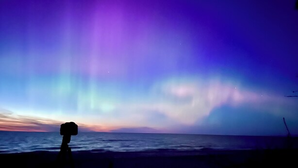 Silueta de una persona frente a una aurora vibrante sobre el océano al anochecer.