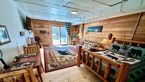 Un acogedor interior de cabaña con piso de madera, un sofá con cojines de osos, una mesa de comedor con sillas y un ventanal con vistas a árboles.