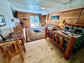 Un acogedor interior de cabaña con piso de madera, un sofá con cojines de osos, una mesa de comedor con sillas y un ventanal con vistas a árboles.