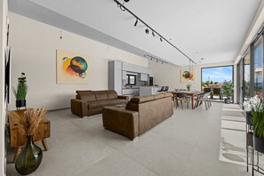 A modern living room with a sofa, coffee table, and a dining area with chairs. There is a kitchen in the background with cabinets and a window.
