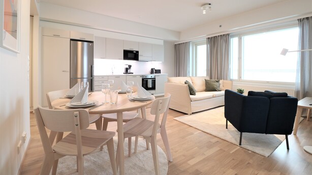 Una sala de estar moderna con un sofá, una mesa de comedor preparada para dos personas y una cocina al fondo.
