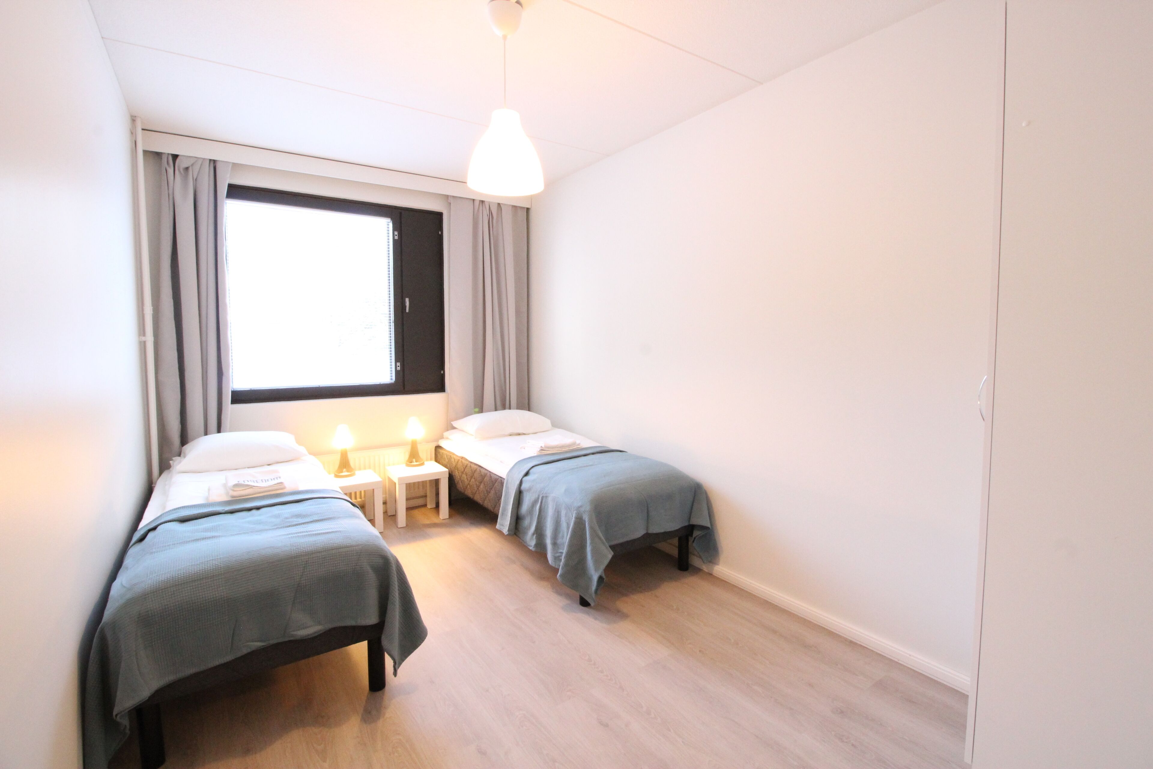 A small, well-lit bedroom with two beds, a window with curtains, a bedside table with a lamp, and a hanging light fixture.