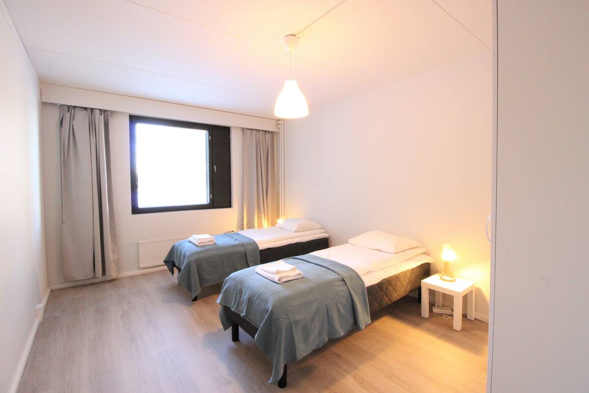 A room with two beds, a window with curtains, a standing lamp, and a radiator.