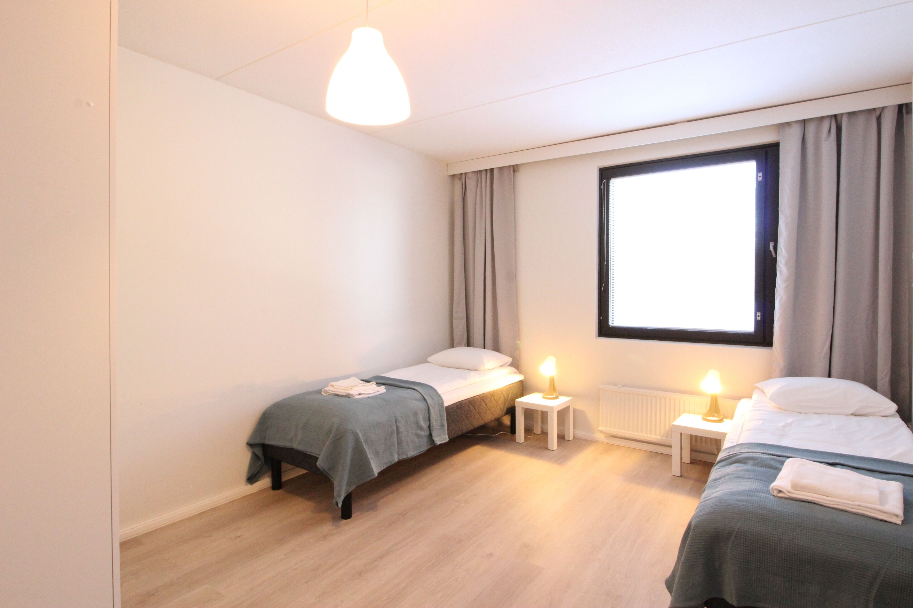 A small, well-lit bedroom with two beds, a window with curtains, a bedside table with a lamp, and a radiator.