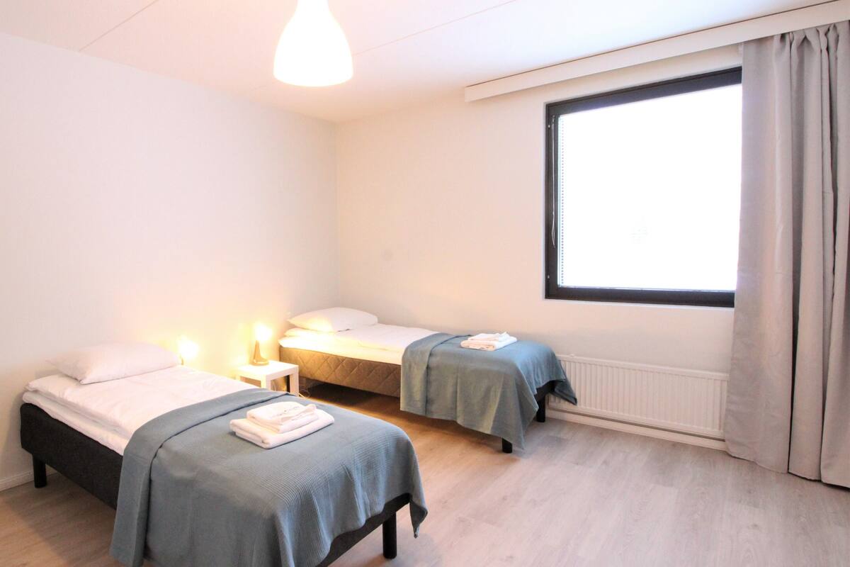 A room with two beds, a window with curtains, a bedside table with a lamp, and a radiator.
