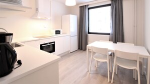 Una cocina moderna con armarios blancos, microondas, lavavajillas y cafetera. Hay una mesa de comedor con sillas y una ventana con cortinas.