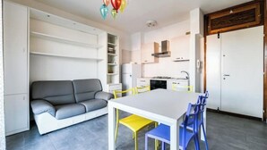 Una sala de estar moderna con un sofá gris, una mesa de comedor blanca con sillas azules y amarillas, y una cocina al fondo.