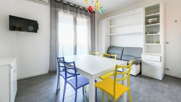 Una sala de estar moderna con un sofá blanco, una mesa de comedor con sillas azules y amarillas, un televisor montado en la pared y una estantería.