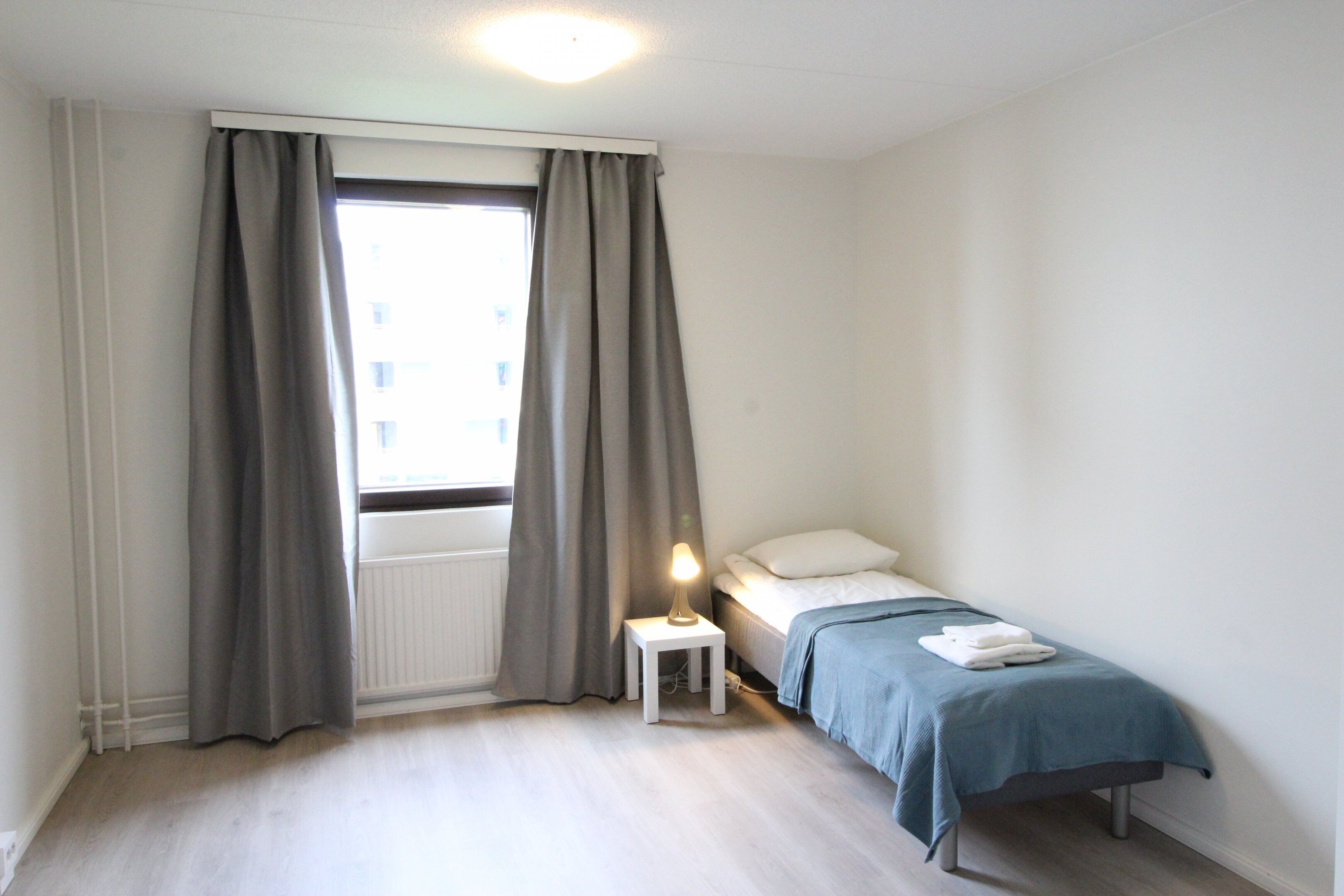 A small, clean room with a single bed, a nightstand with a lamp, and a window with curtains.