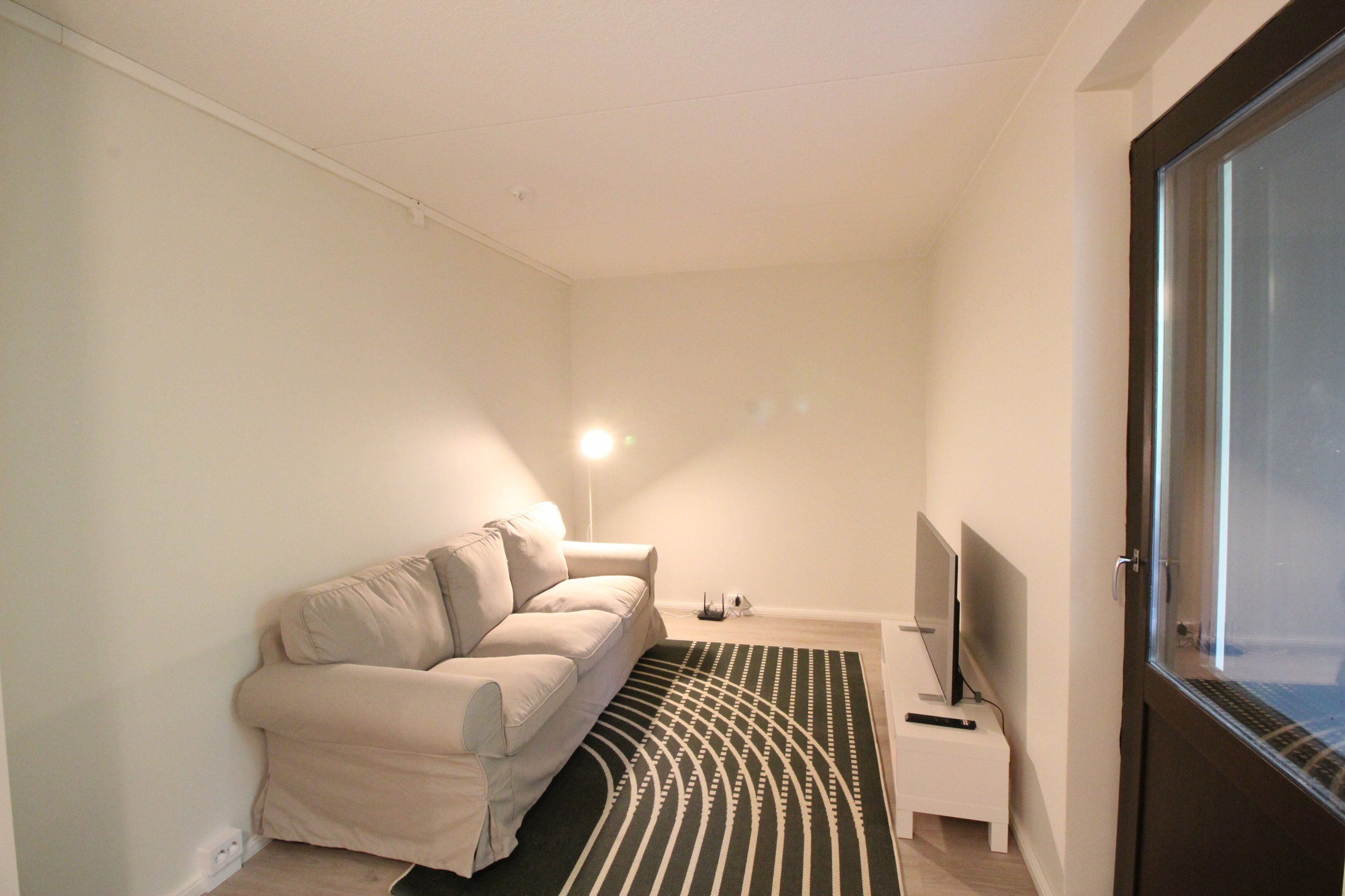 A small, modern living room with a sofa, a flat-screen TV, a lamp, and a window.