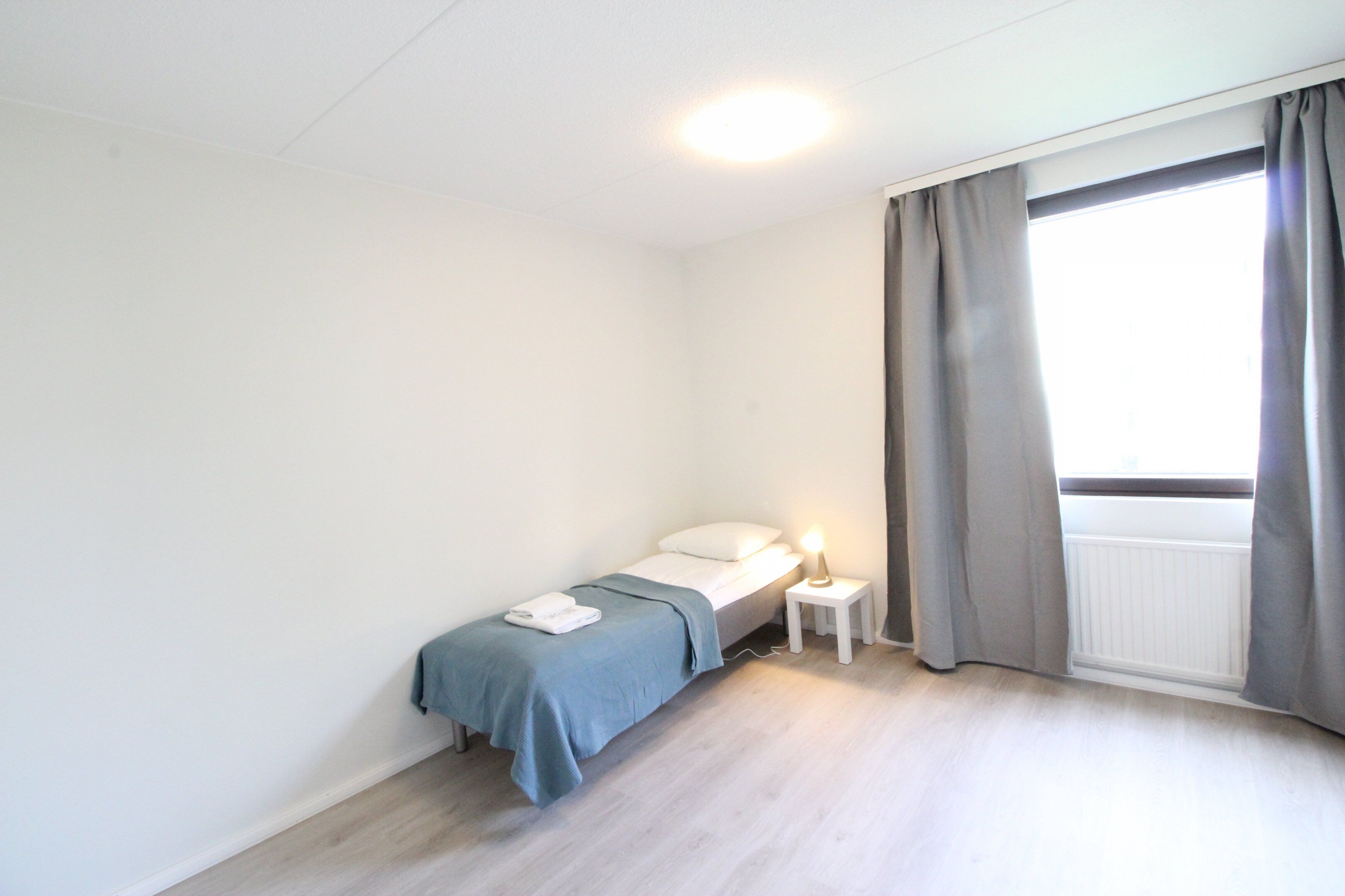 A small, clean room with a bed, bedside table, and a window with curtains.