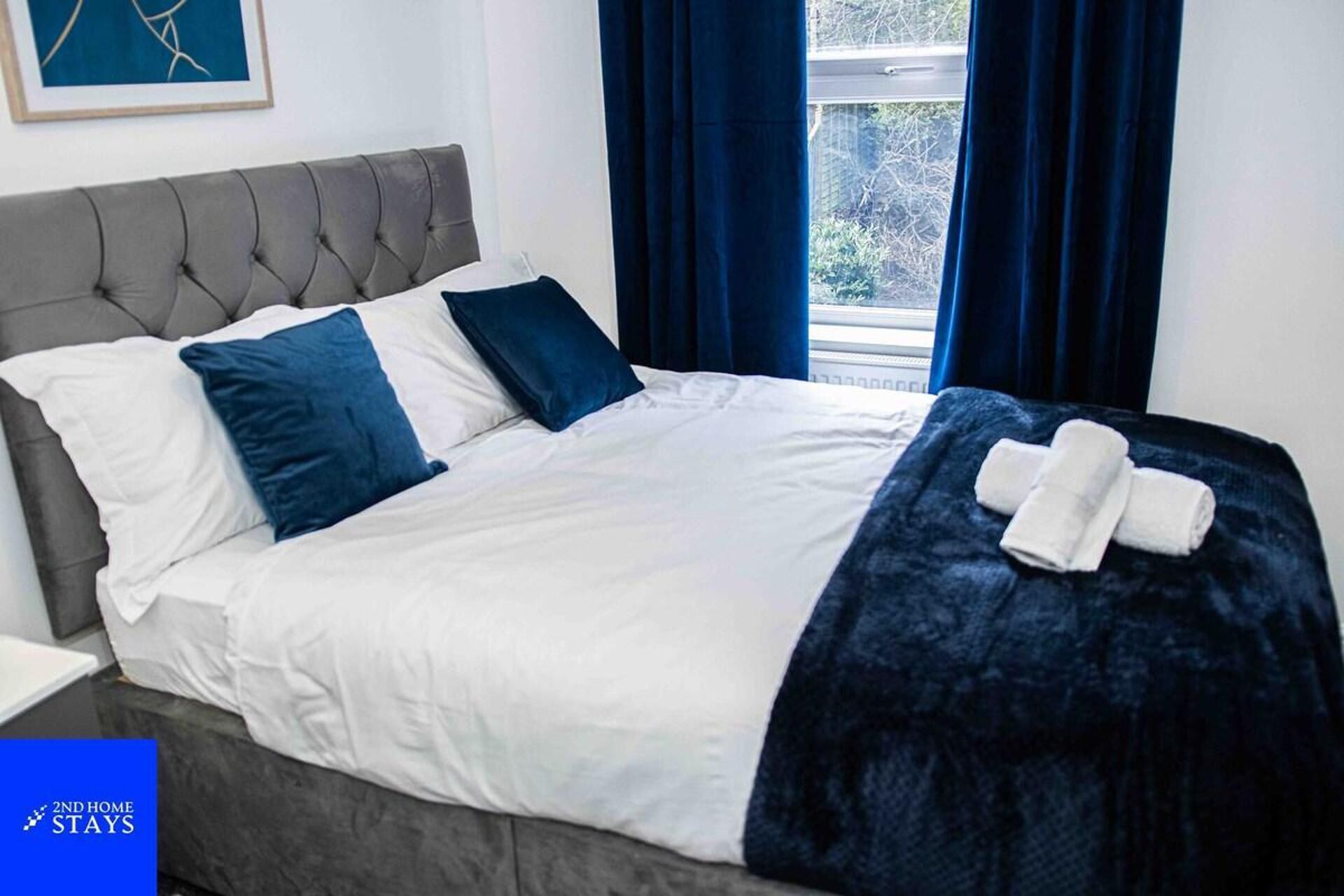 Een net opgemaakt bed met wit- en blauwe beddengoed, een grijs gestoffeerd hoofdbord en een donkerblauwe plaid, met handdoeken aan de zijkant.