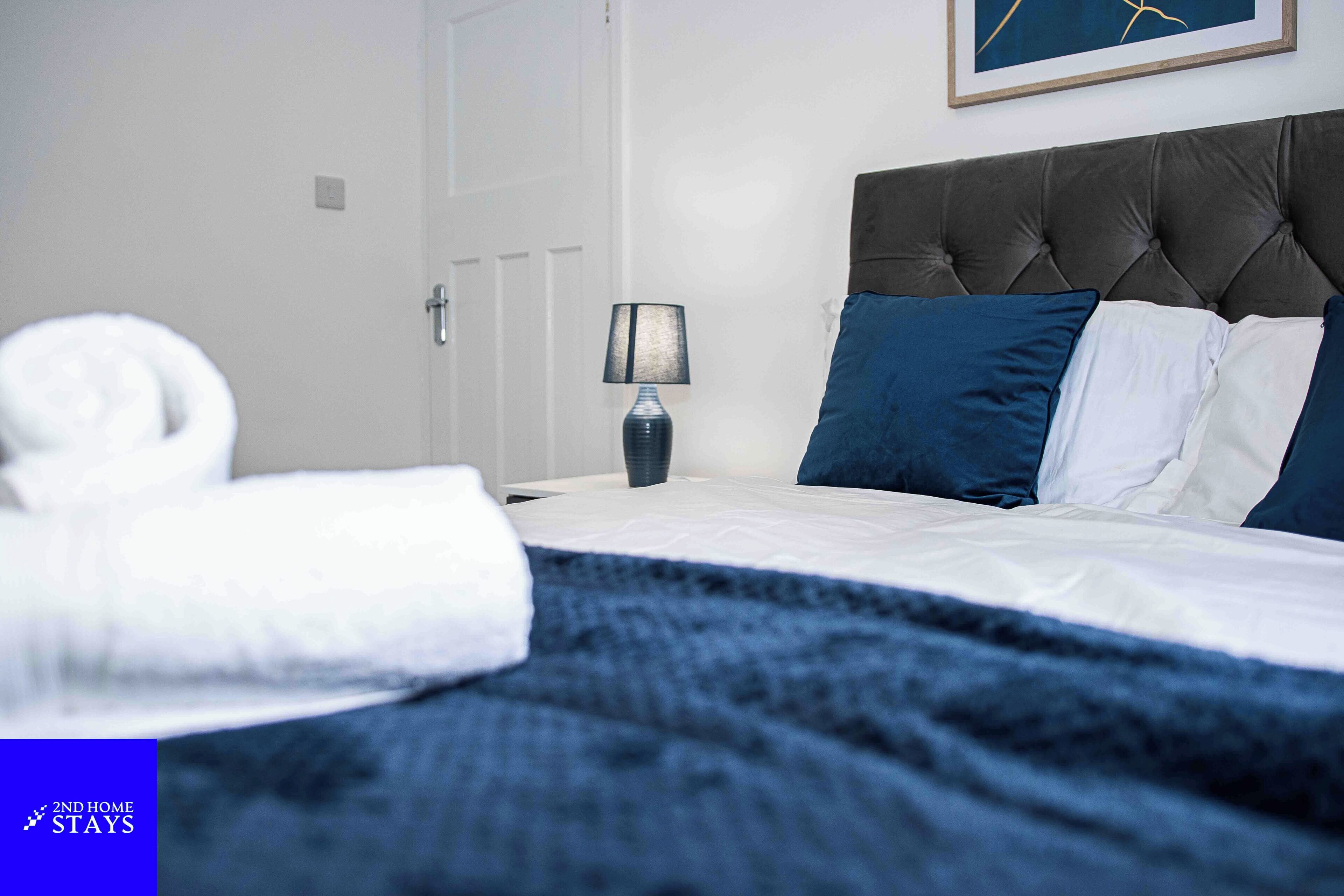 Una cama bien hecha con un edredón azul oscuro, una almohada blanca y una almohada azul. Una toalla blanca está en el suelo junto a la cama. Una lámpara con pantalla azul está sobre la mesita de noche. La habitación tiene una puerta blanca y un cuadro enmarcado en la pared sobre la cama.