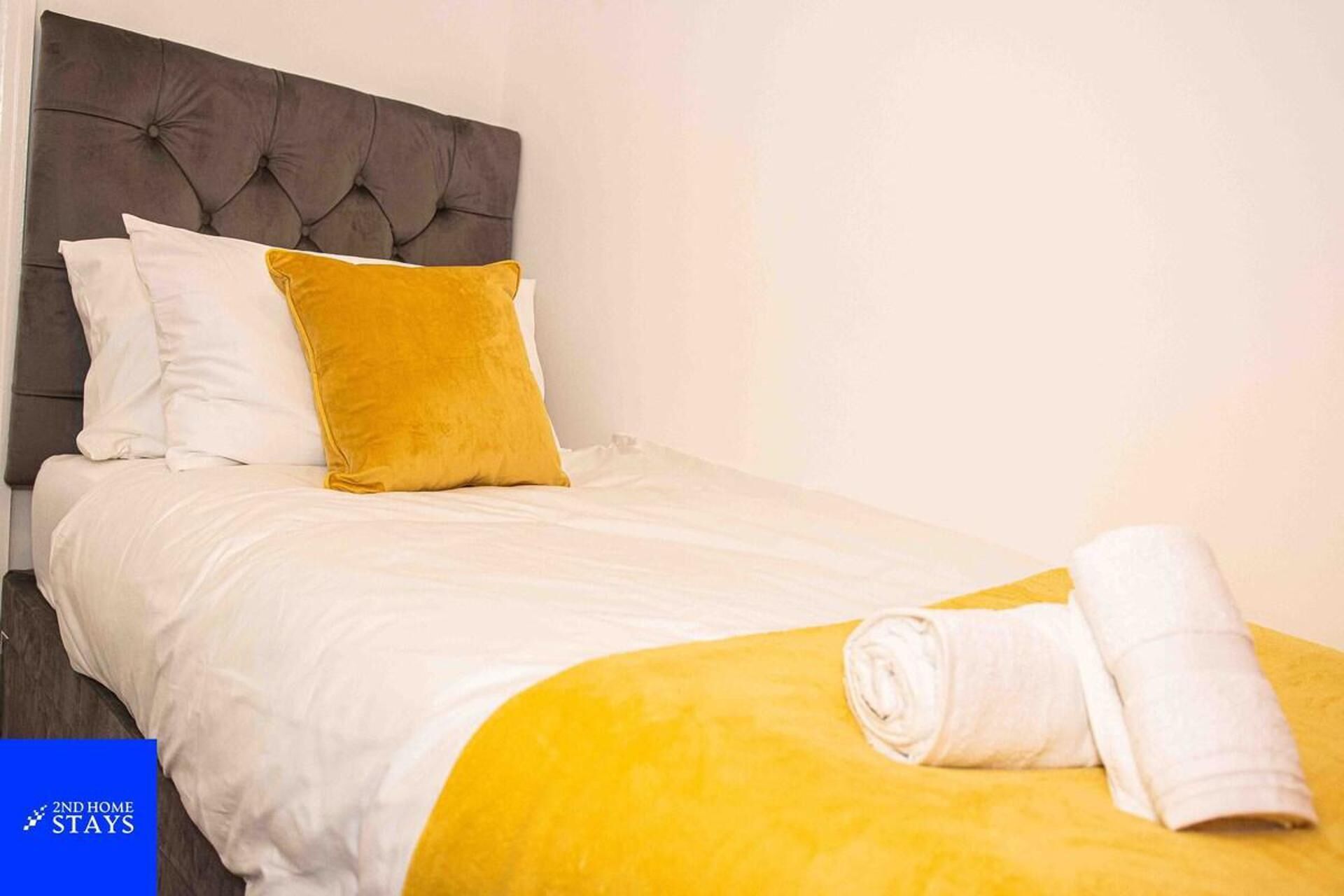 A neatly made bed with a grey headboard, white and yellow pillows, and a yellow blanket. A rolled white towel is placed at the foot of the bed.