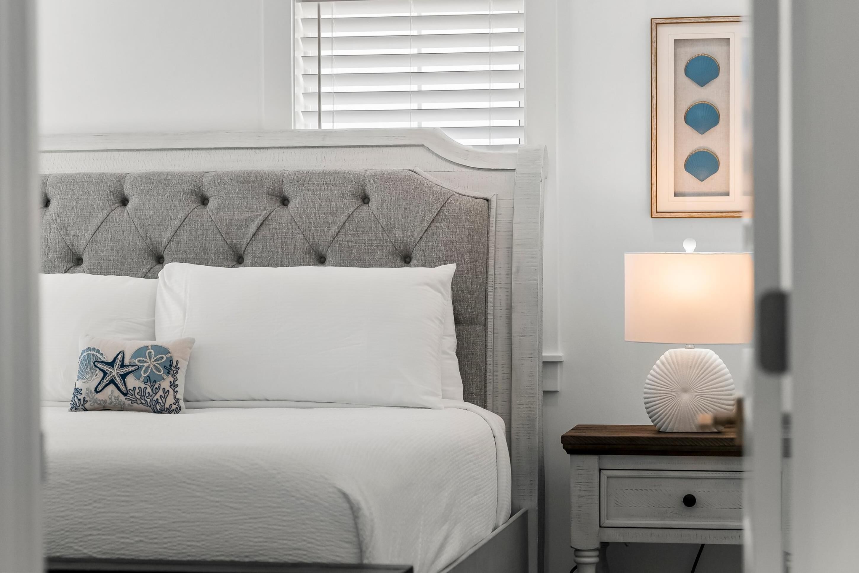 Een netjes opgemaakt bed met een grijs gestoffeerd hoofdbord, een wit kussen en een decoratief kussen met een ster- en koraalmotief. Een nachtkastje met een lamp en een ingelijste foto hangen aan de muur.