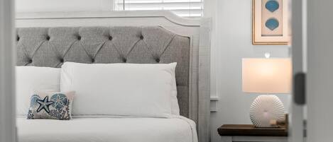 Een netjes opgemaakt bed met een grijs gestoffeerd hoofdbord, een wit kussen en een decoratief kussen met een ster- en koraalmotief. Een nachtkastje met een lamp en een ingelijste foto hangen aan de muur.