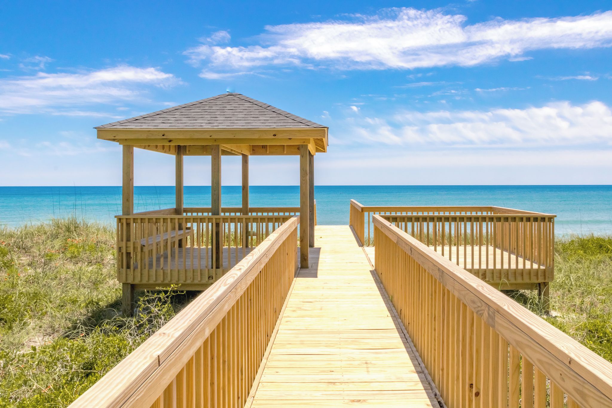 A wooden gazebo with a roof, connected by a wooden walkway leading to a beach.