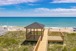 Une structure en bois installée sur la plage, dotée d’un toit et entourée de chaises de plage et de parasols, offrant une vue sur une plage de sable et une mer calme.