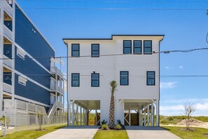 A two-story building with a covered entrance and a palm tree in the foreground. - Shore N' Tell - Luxury 5BR Home - Cov. Porch, Elevator, Gazebo and More! (Surf City)