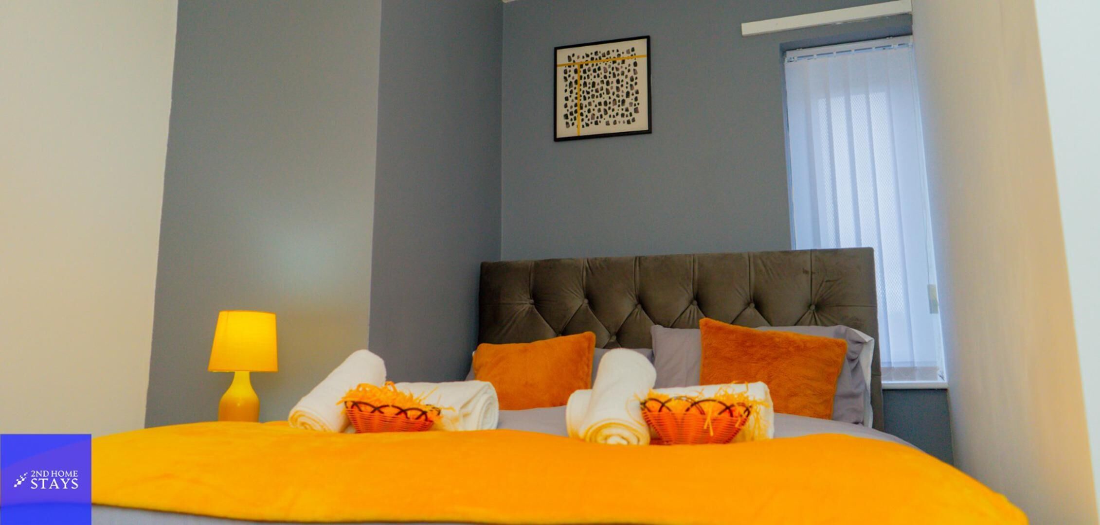 Habitación de hotel con un cabecero capitonado, una cama con ropa de cama naranja, una lámpara de noche y una ventana con persianas.