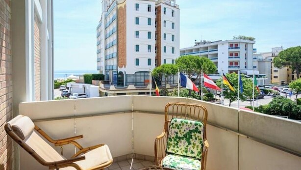 Un balcon avec deux fauteuils en osier, une vue sur un paysage urbain avec des bâtiments et des drapeaux, et un ciel dégagé.