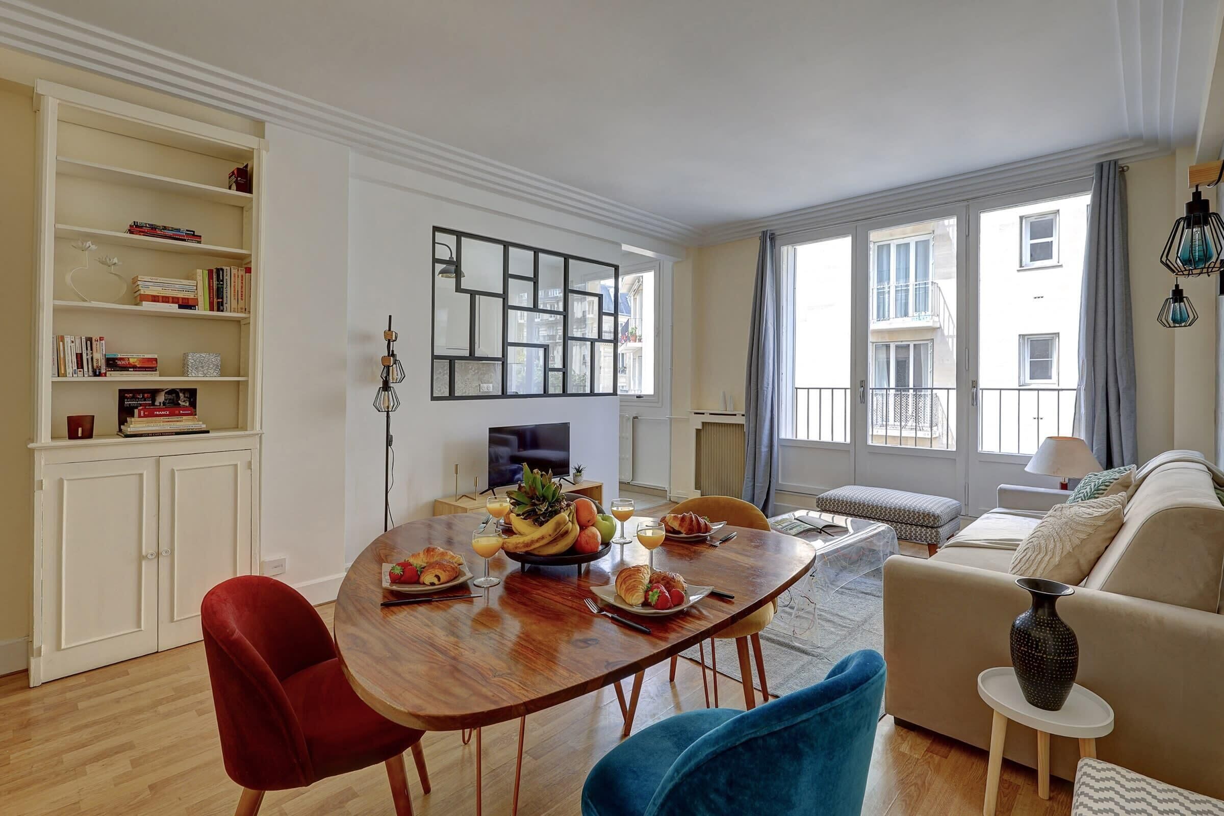 A modern living room with a wooden dining table, red and blue chairs, a beige sofa, and a small side table with a vase.
