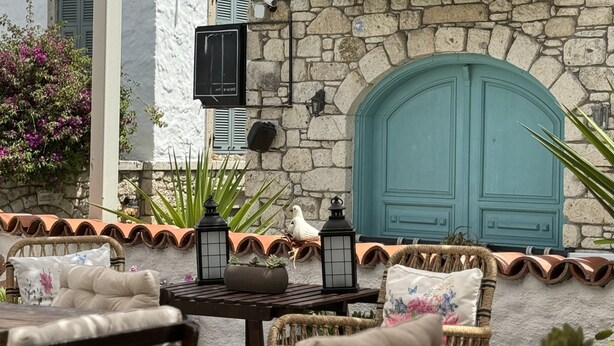 An outdoor seating area with wicker chairs and a table, a stone wall with a teal door, and a white building with shutters. - H&R Alaçatı Hotel (Cesme)
